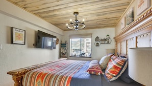A bedroom with a wooden ceiling, a bed with a colorful blanket, a mounted TV, and a window with a view of buildings. - Ski-in Red River Retreat: Balcony, Mountain Views! (Red River)