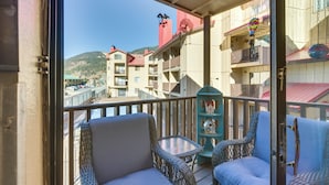 A balcony with wicker chairs and a small table, overlooking a waterfront with buildings and a mountain in the background. - Ski-in Red River Retreat: Balcony, Mountain Views! (Red River)