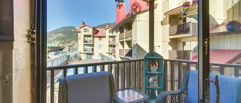 A balcony with wicker chairs and a small table, overlooking a waterfront with buildings and a mountain in the background.