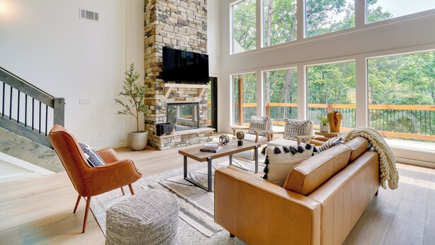 A spacious living room with a stone fireplace, large windows, and a wooden staircase.