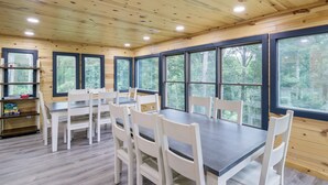 A wooden cabin interior with a dining table and chairs, large windows, and a bookshelf.