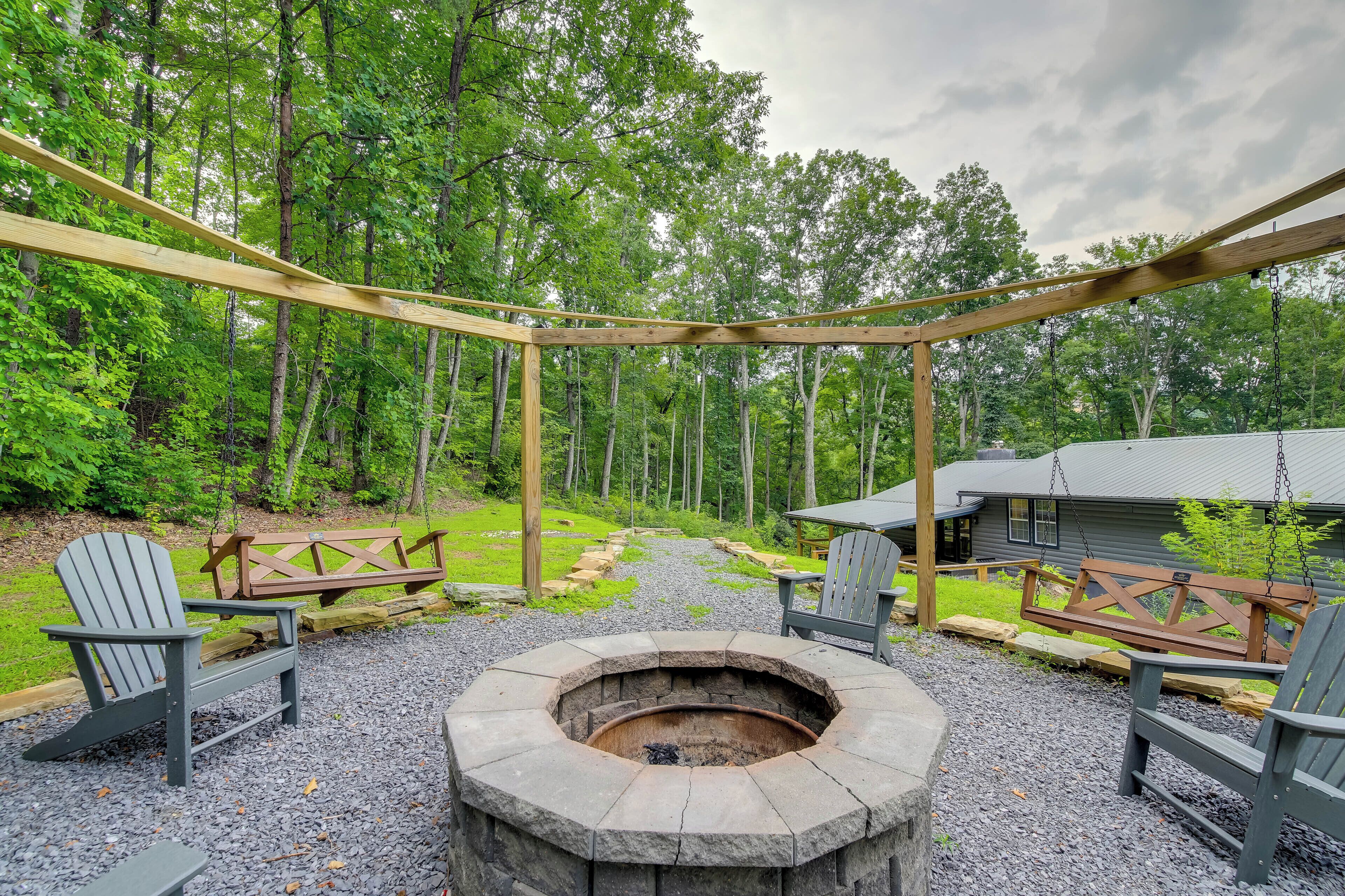 Un área de estar al aire libre rústica con sillas Adirondack y un hogar de piedra, rodeada por una pérgola de madera y un bosque de fondo.