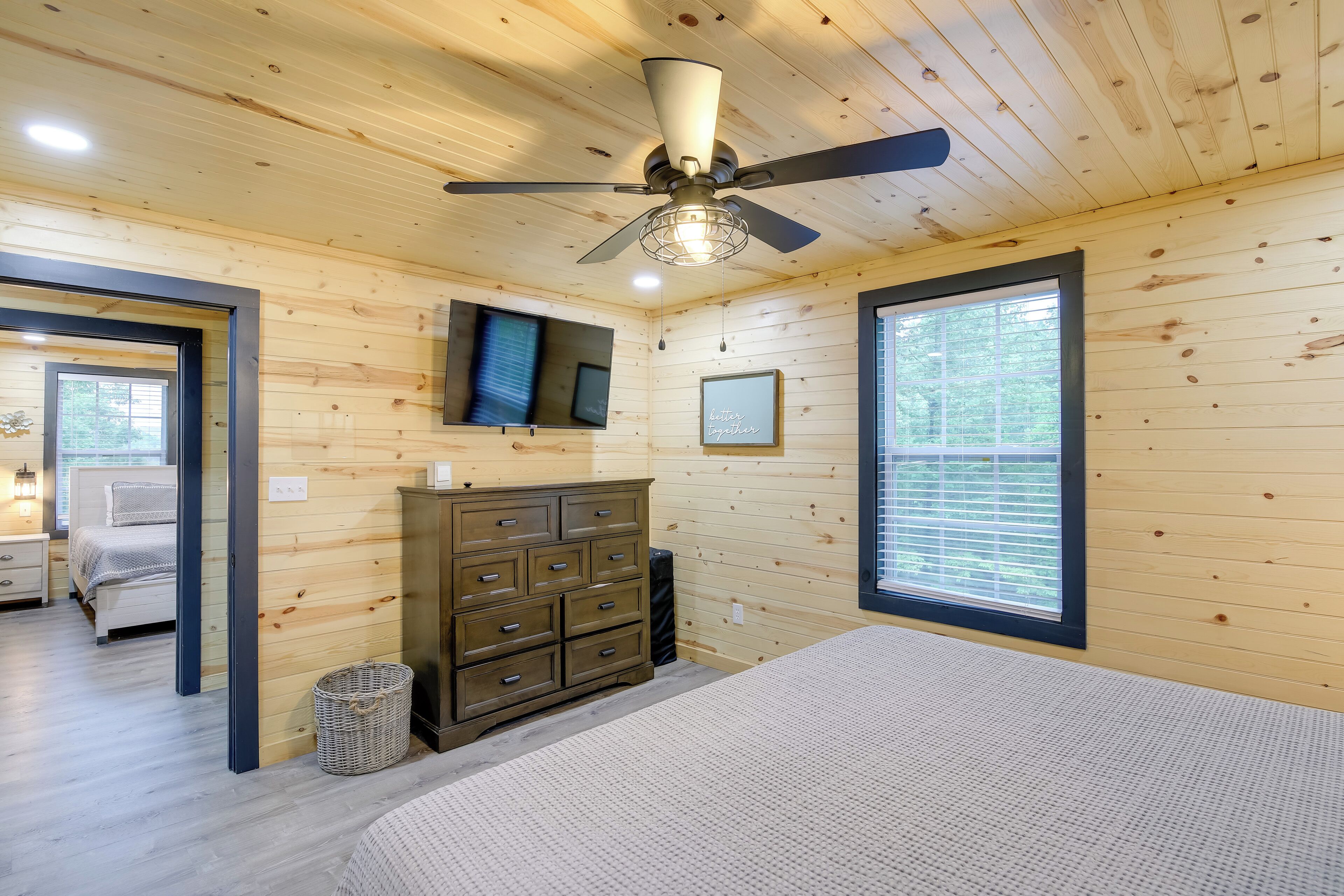 Un dormitorio con paredes de madera, ventilador de techo, cama, cómoda, televisor y ventana.