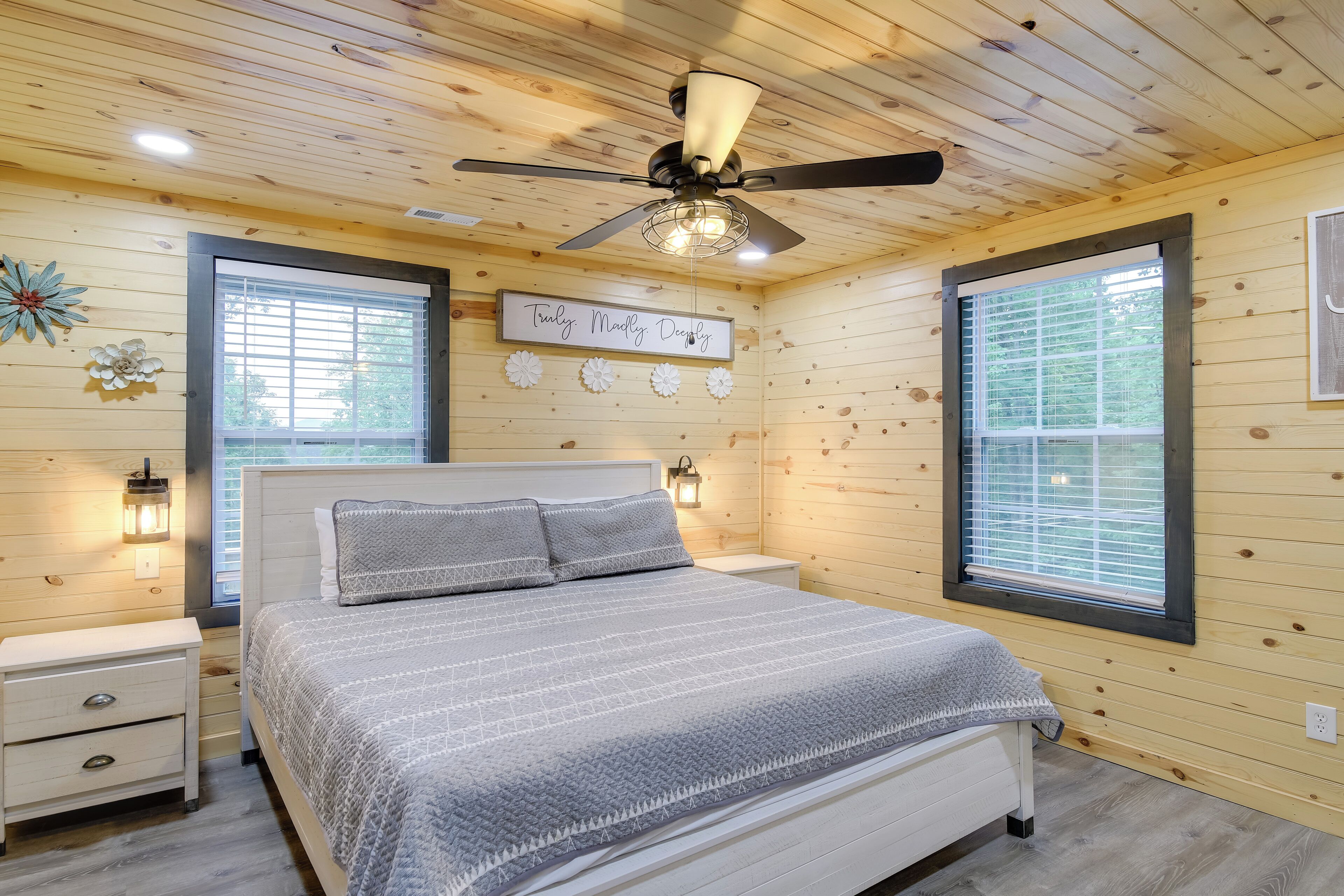 Ein Schlafzimmer mit Bett, Nachttischen, Deckenventilator und zwei Fenstern mit Jalousien.