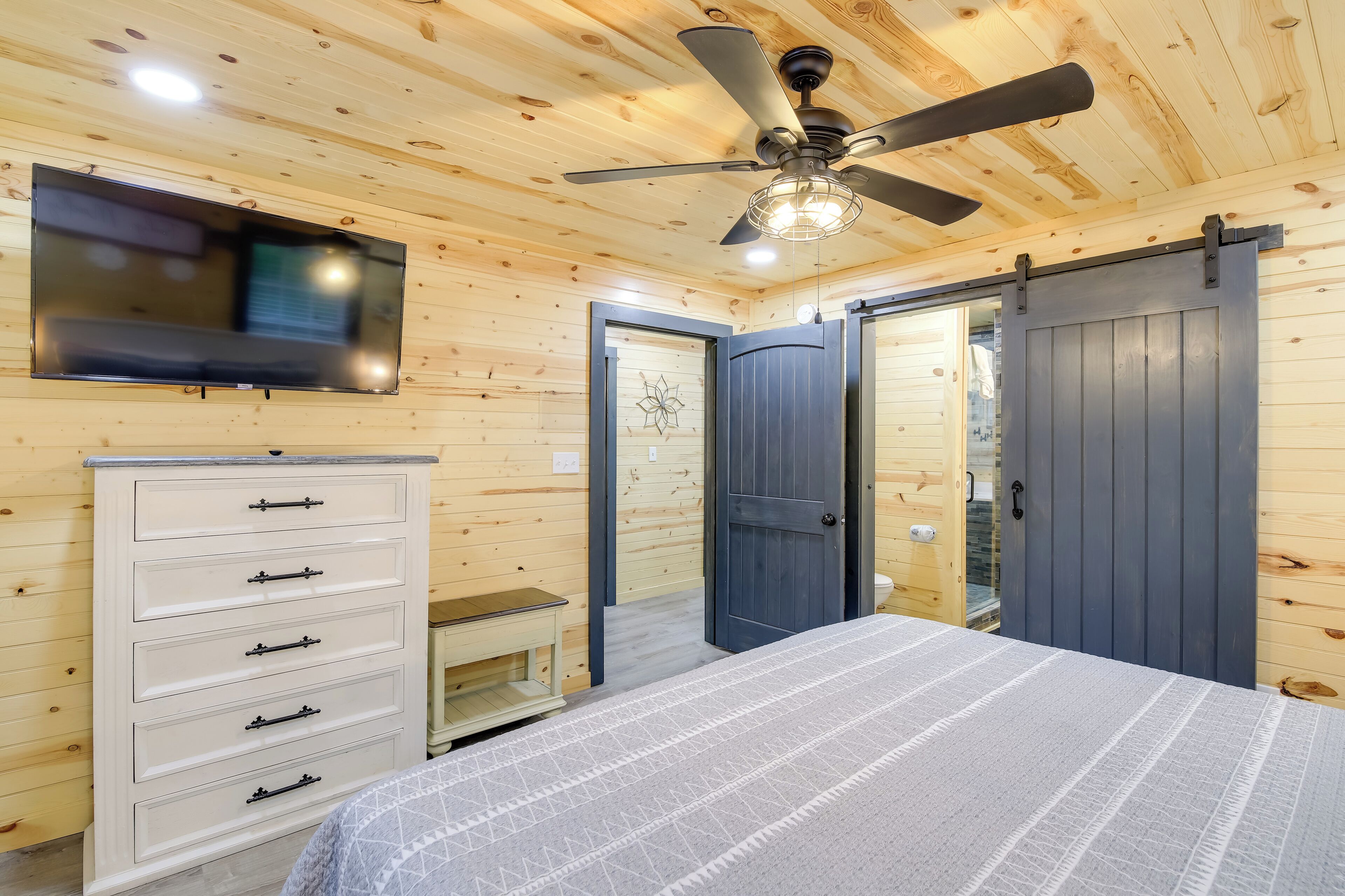 A bedroom with a bed, a dresser, a ceiling fan, and a TV mounted on the wall.