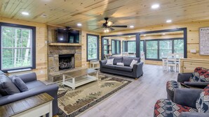 A cozy cabin interior with a fireplace, a ceiling fan, and large windows offering a view of the forest.
