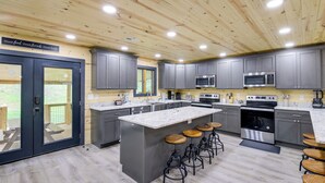 A modern kitchen with grey cabinets, a large island with stools, stainless steel appliances, and a wooden ceiling.