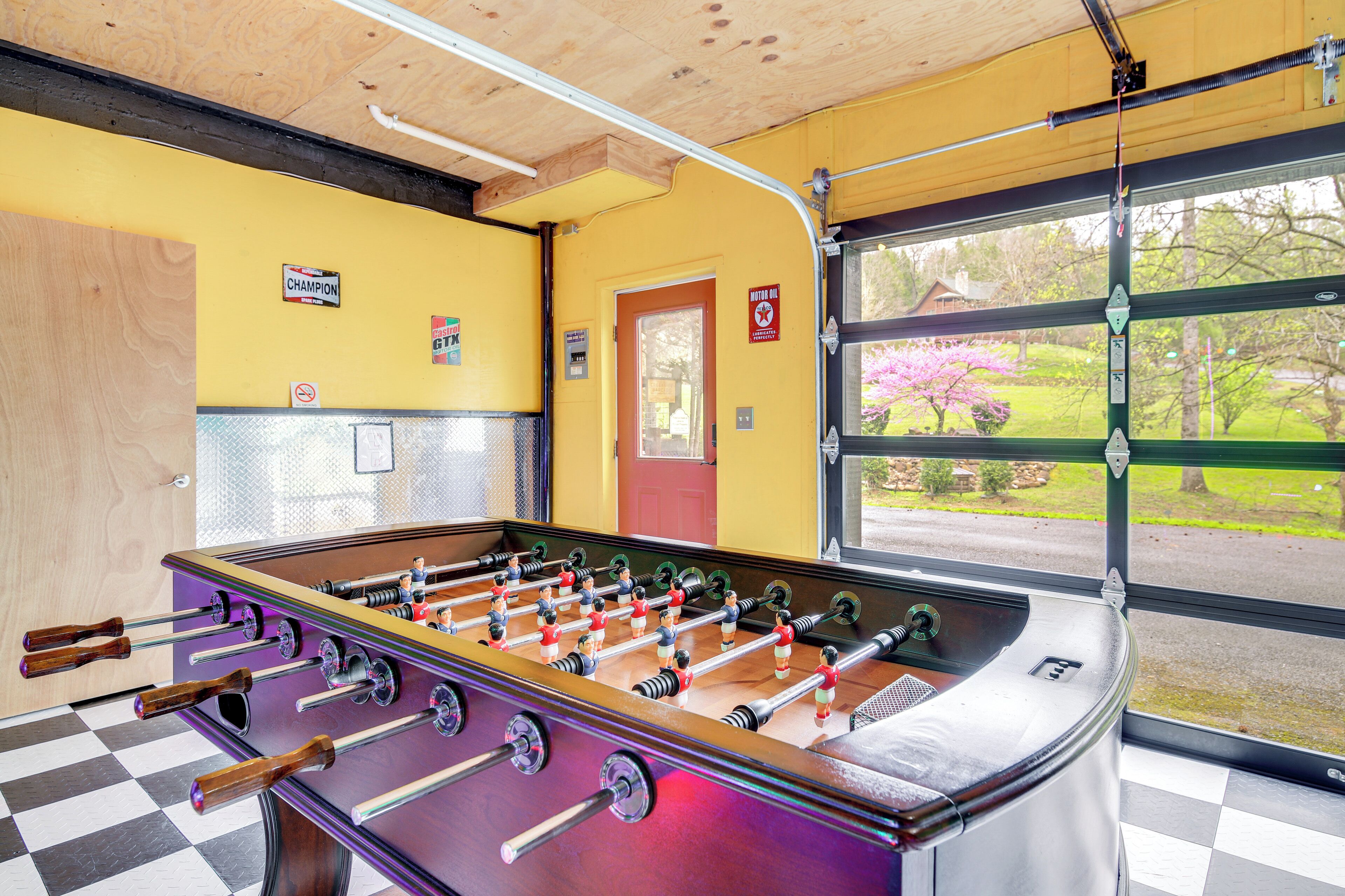 A foosball table in a room with a checkered floor, yellow walls, and a window with a view of a garden.