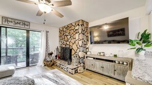 A cozy living room with a stone fireplace, a ceiling fan, a large window, and a wooden dining table. - Sevierville Condo w/ Balcony + Pool Access! (Sevierville)