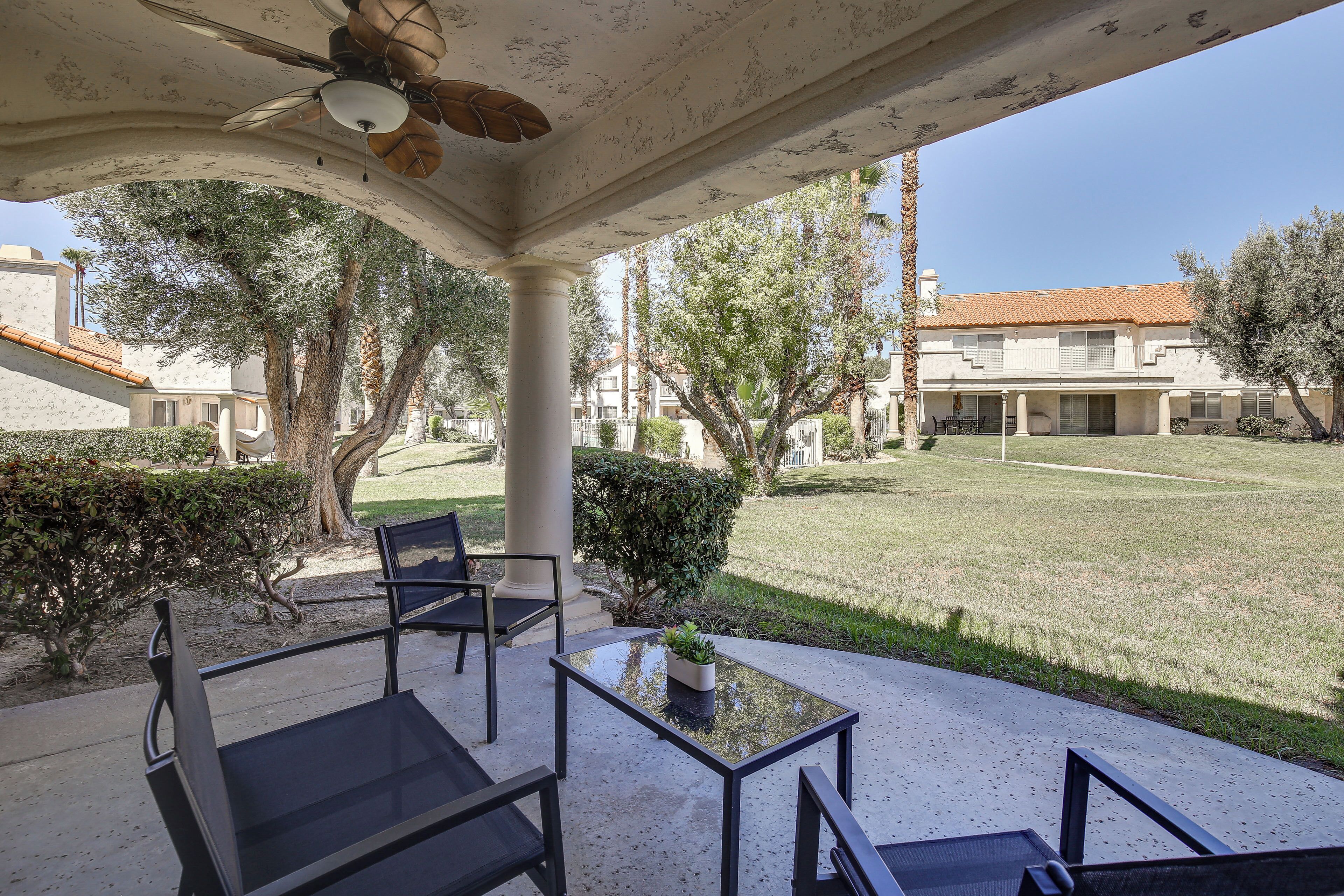 A covered outdoor seating area with a table and chairs, a ceiling fan, and a view of a building and lawn.