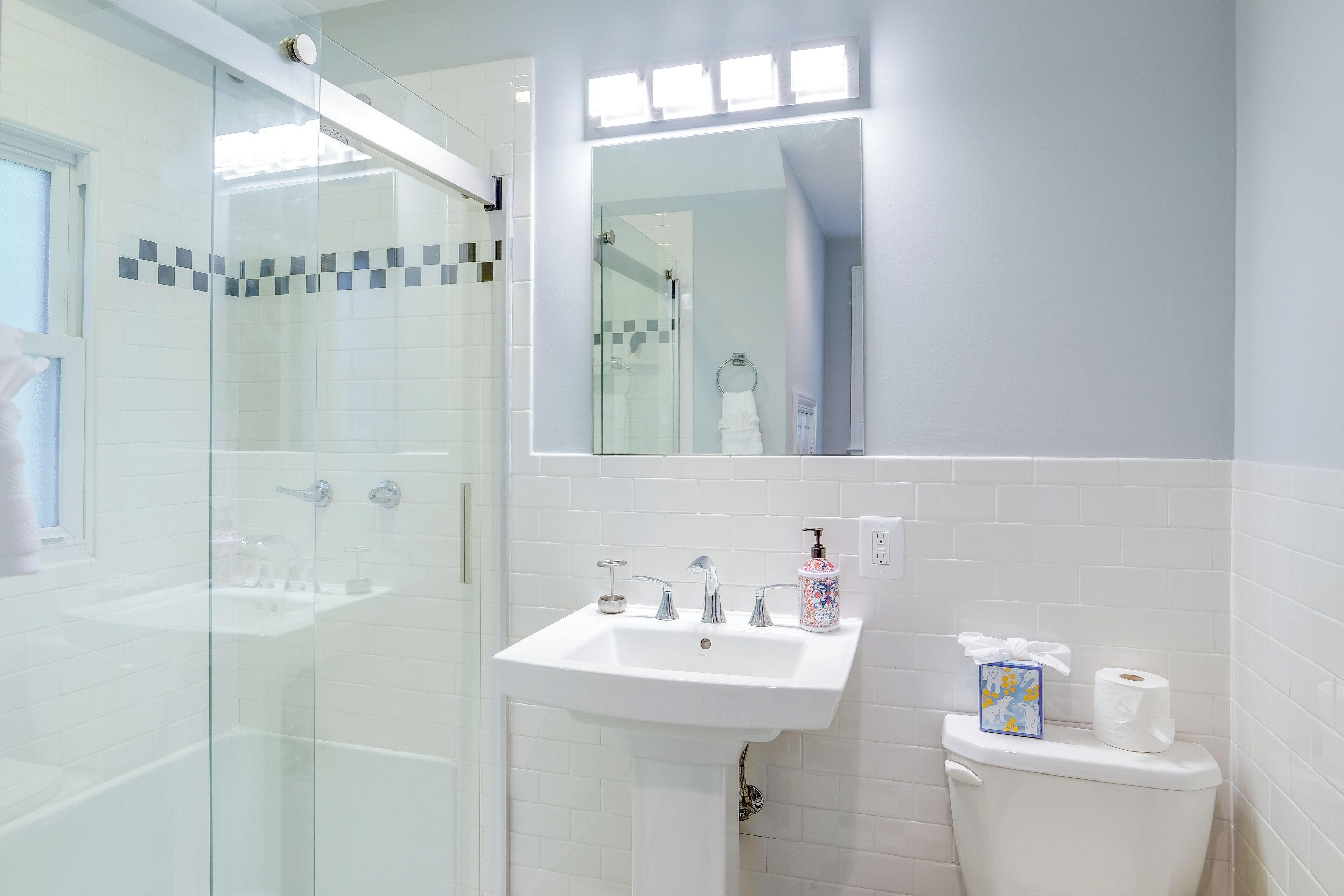 A bathroom with a glass shower enclosure, a white sink with two faucets, and a toilet with a tissue box and toilet paper.