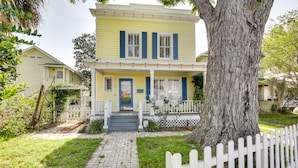 Une maison à deux étages avec un porche à l’avant, une clôture en bois peinte en blanc et un grand arbre au premier plan.