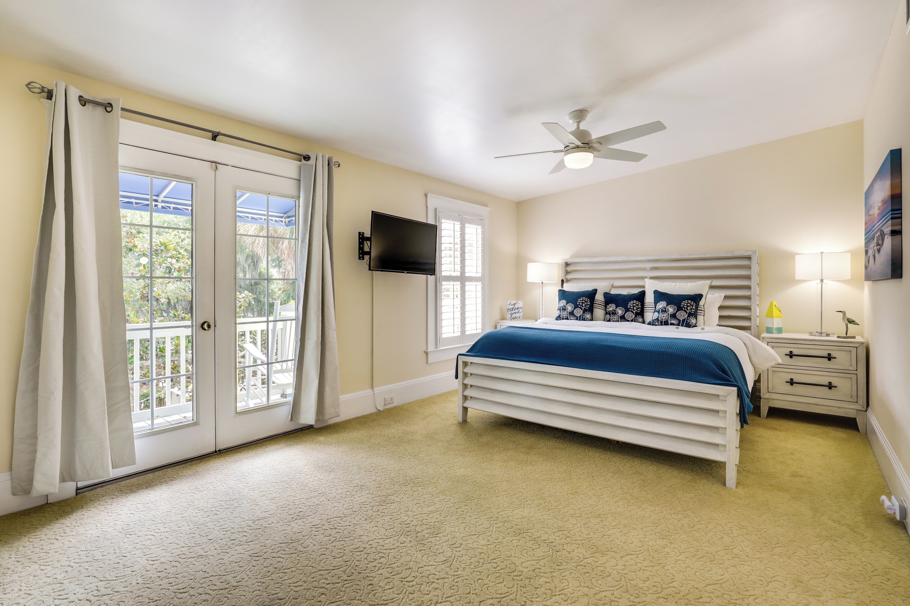 Un dormitorio con cama, ventilador de techo, televisor y balcón con cortinas.
