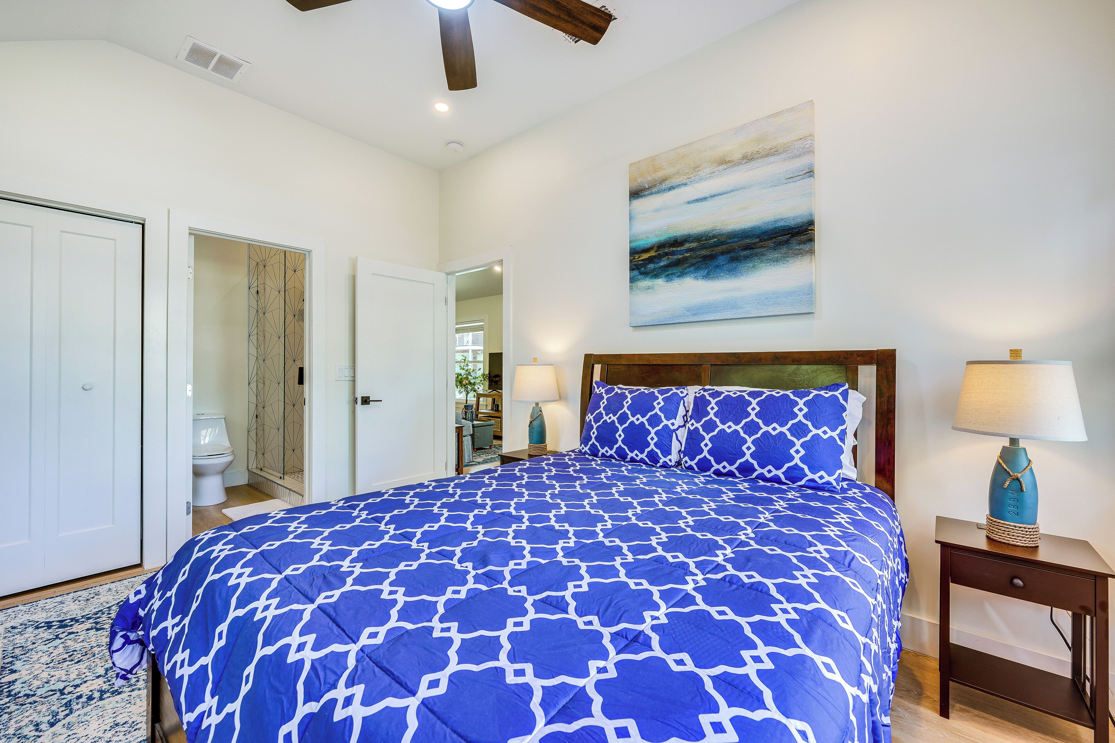 A bedroom with a large bed, a nightstand, a lamp, a painting on the wall, and a view into the adjacent room.