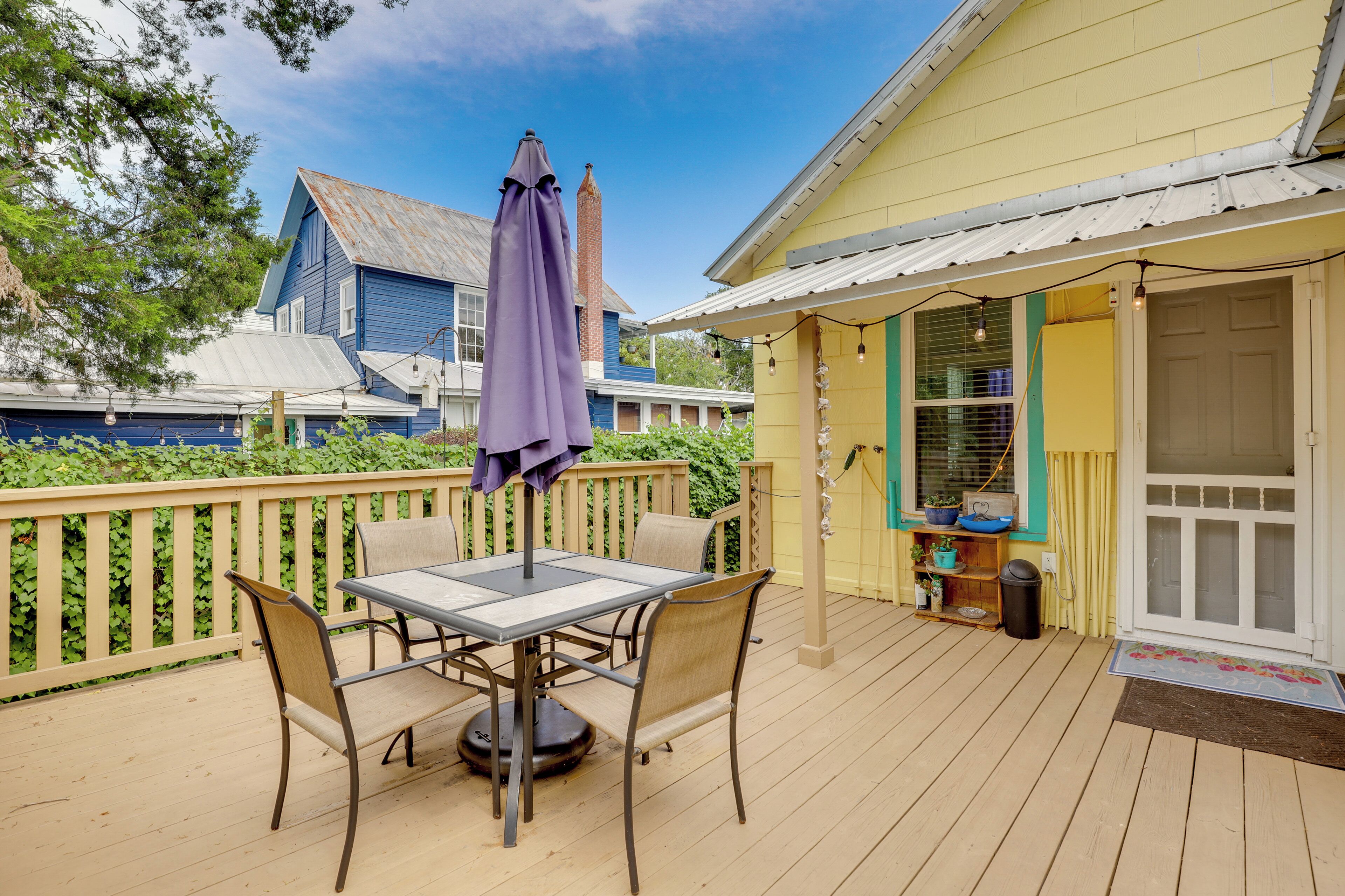 Eine Holzterrasse mit Tisch und Stühlen, einem violetten Sonnenschirm und einem gelben Gebäude mit Fenster und Tür.