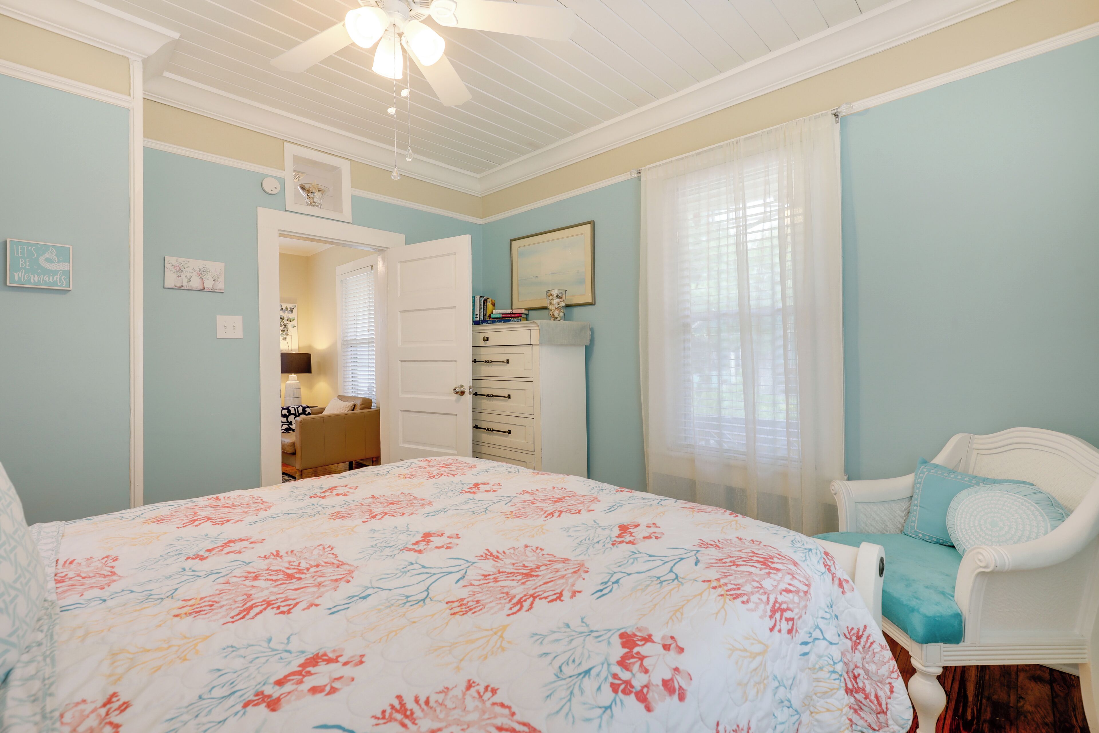 Quarto com colcha florida, penteadeira branca, cadeira com almofada azul e ventilador de teto.