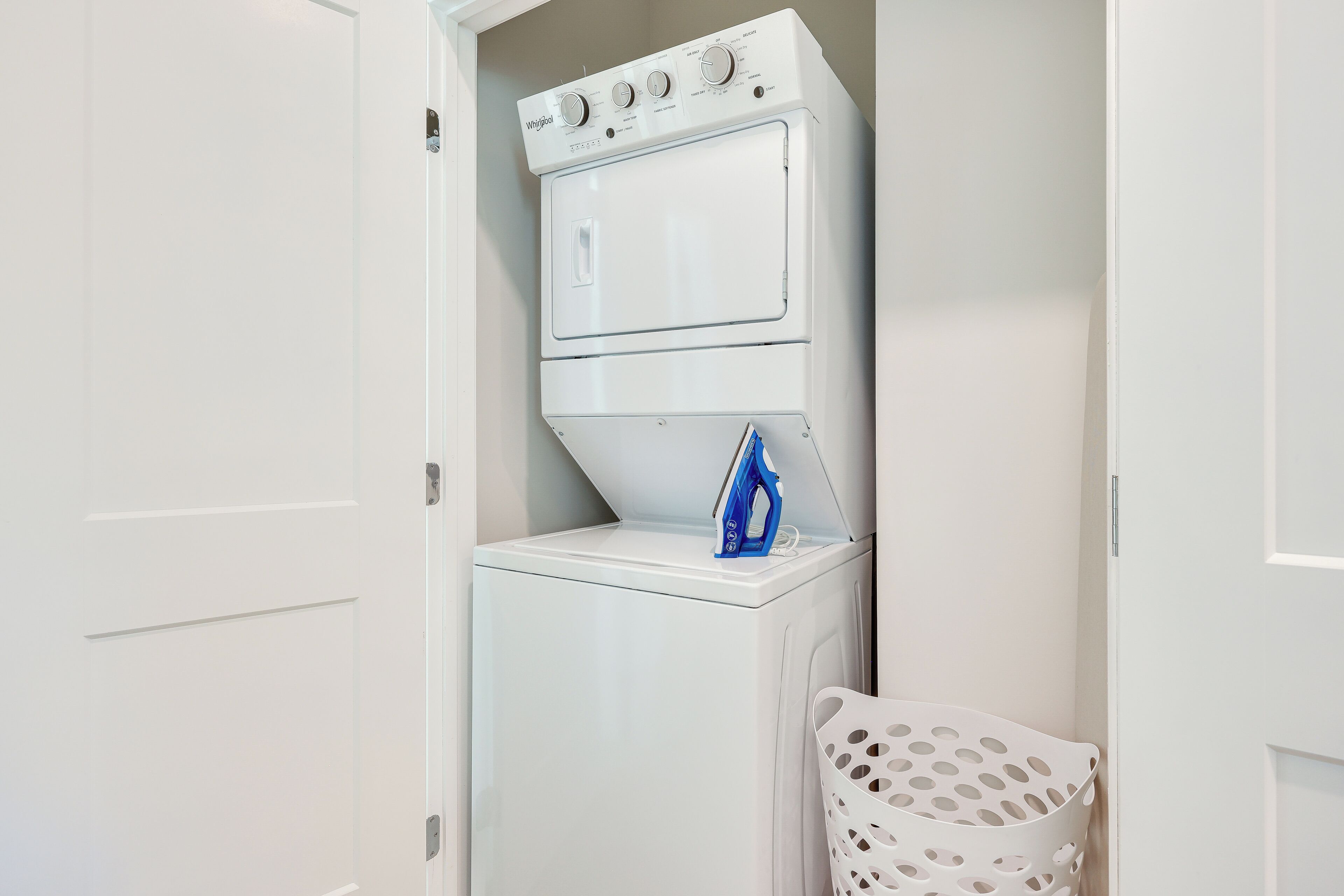 A compact laundry area with a stacked washer and dryer, a blue iron, and a white laundry basket.