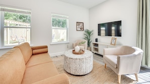 A modern living room with a beige sofa, a round coffee table, a white armchair, and a flat-screen TV mounted on the wall. - St Augustine Home < 3 Mi to Historic District! (St. Augustine)