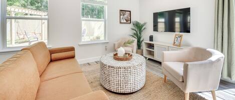 A modern living room with a beige sofa, a round coffee table, a white armchair, and a flat-screen TV mounted on the wall.