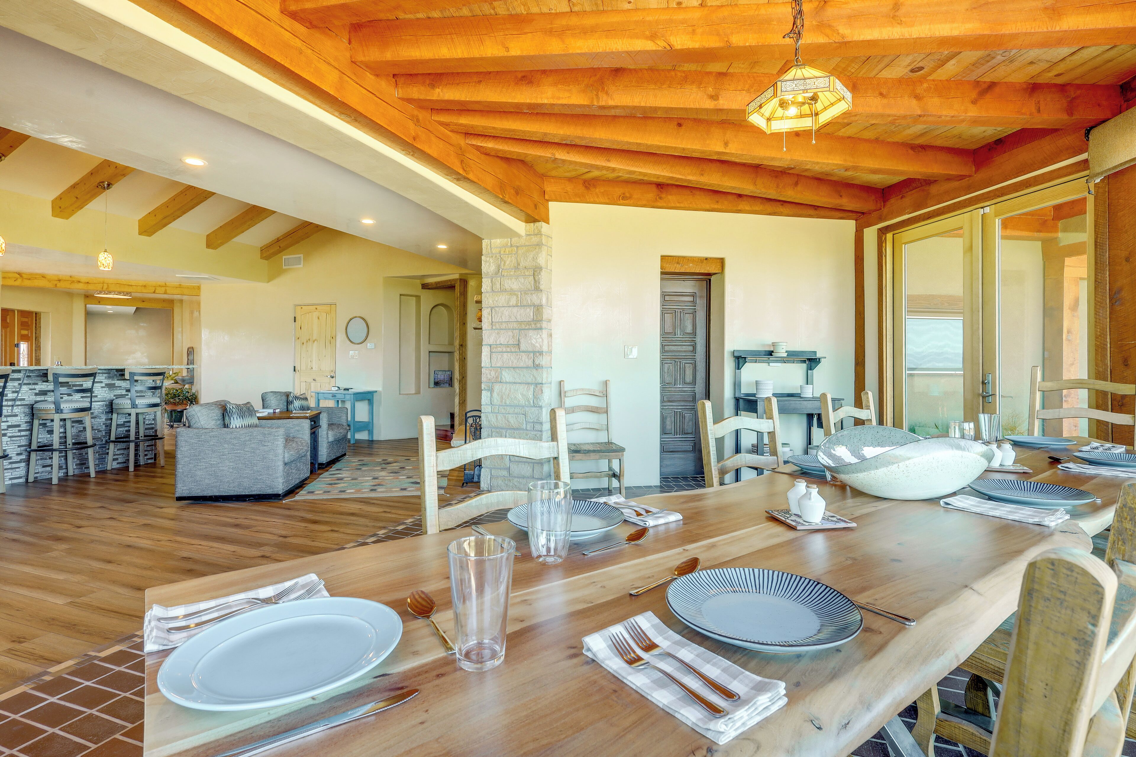 Un comedor con una mesa de madera dispuesta para cuatro personas, rodeada de sillas. Se observa una pared de piedra y una puerta al fondo.