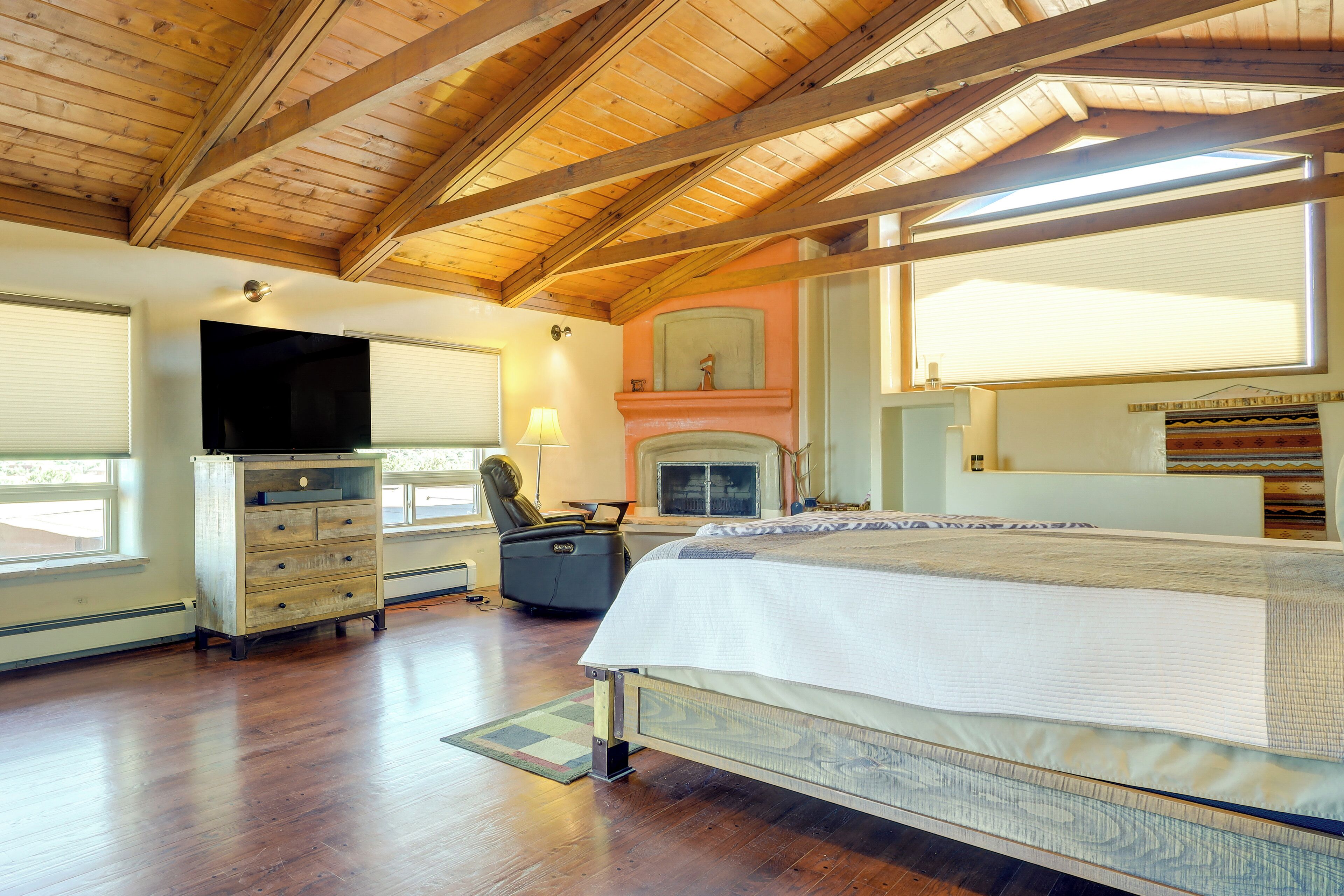 A bedroom with a large bed, wooden ceiling beams, a fireplace, a television, and a dresser.
