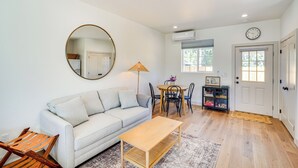 A living room with a sofa, a coffee table, a dining table with chairs, a lamp, a bookshelf, and a door with a window. - Modern Studio in Bend ~ 3 Mi to Sunriver Resort! (Bend)
