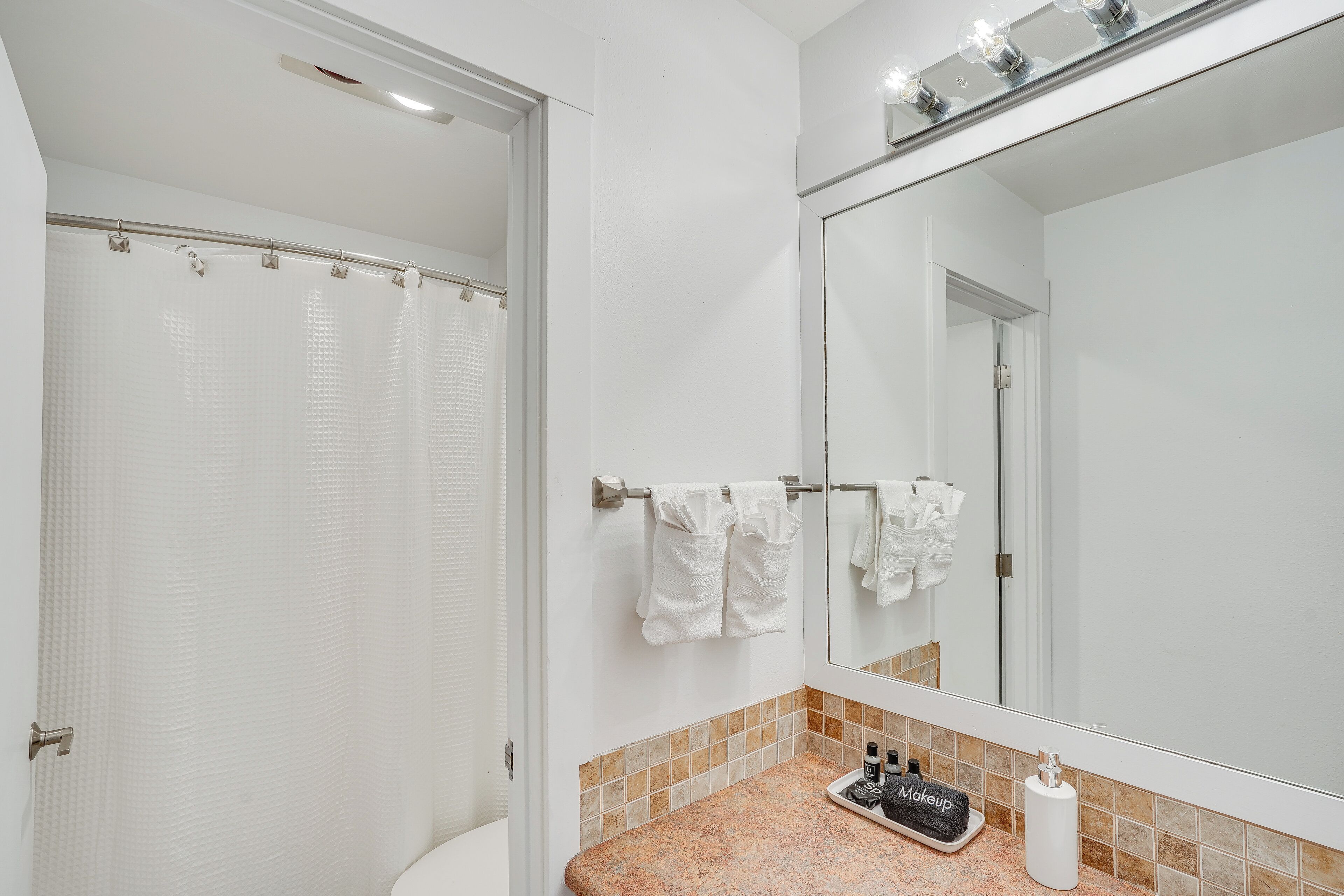 Un baño con cortina de ducha, toallero con toallas, lavabo con espejo y artículos de aseo en la encimera.