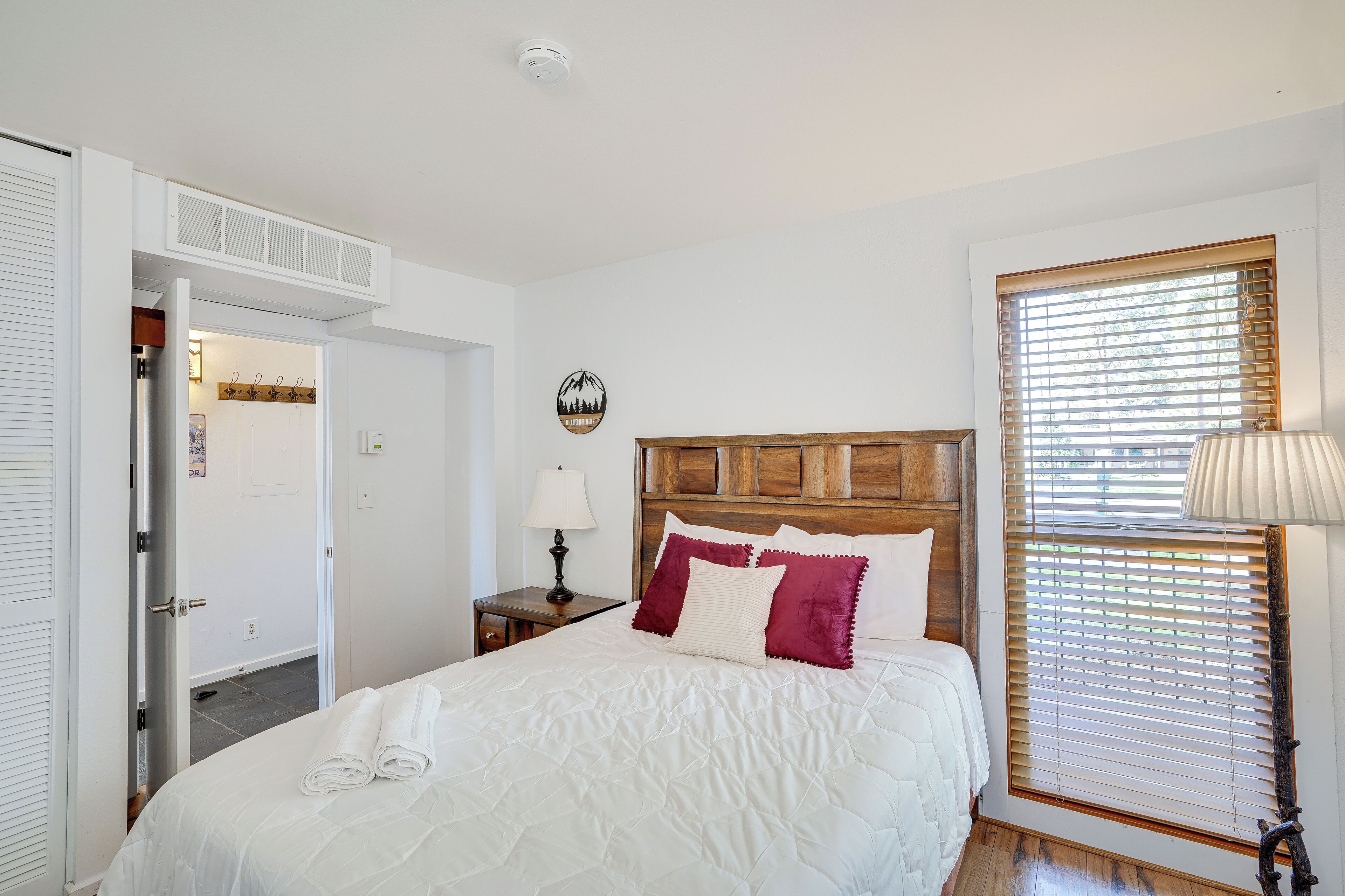 A bedroom with a bed, a nightstand, a lamp, a window with blinds, and a door leading to another room.