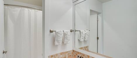 Un baño con cortina de ducha, toallero con toallas, lavamanos con dispensador de jabón y un espejo.