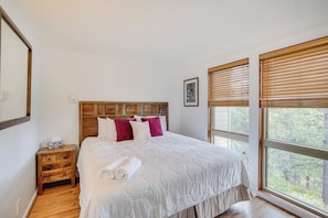 A bedroom with a wooden headboard, a large window with blinds, a bedside table, and a neatly made bed with white bedding and a rolled towel. - Seventh Mountain Condo in Bend w/ Scenic Views! (Bend)