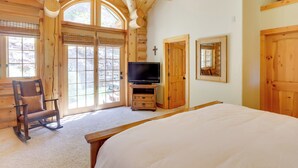 Un dormitorio de cabaña de madera con una cama grande, un televisor sobre un soporte de madera, una mecedora y una pared de piedra visible a través de la puerta.