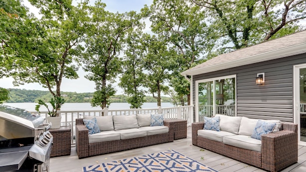 A covered outdoor seating area with wicker sofas and a patterned rug, overlooking a body of water and surrounded by trees. - Dock & Sunset Views: Lakeside Ozark Retreat! (Lake Ozark)