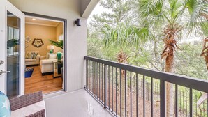 A balcony with a view of a garden, a door leading to a living room with a sofa and a lamp, and a small table with a plant. - Miramar Beach Condo w/ Pool - Walk to Beach! (Miramar Beach)