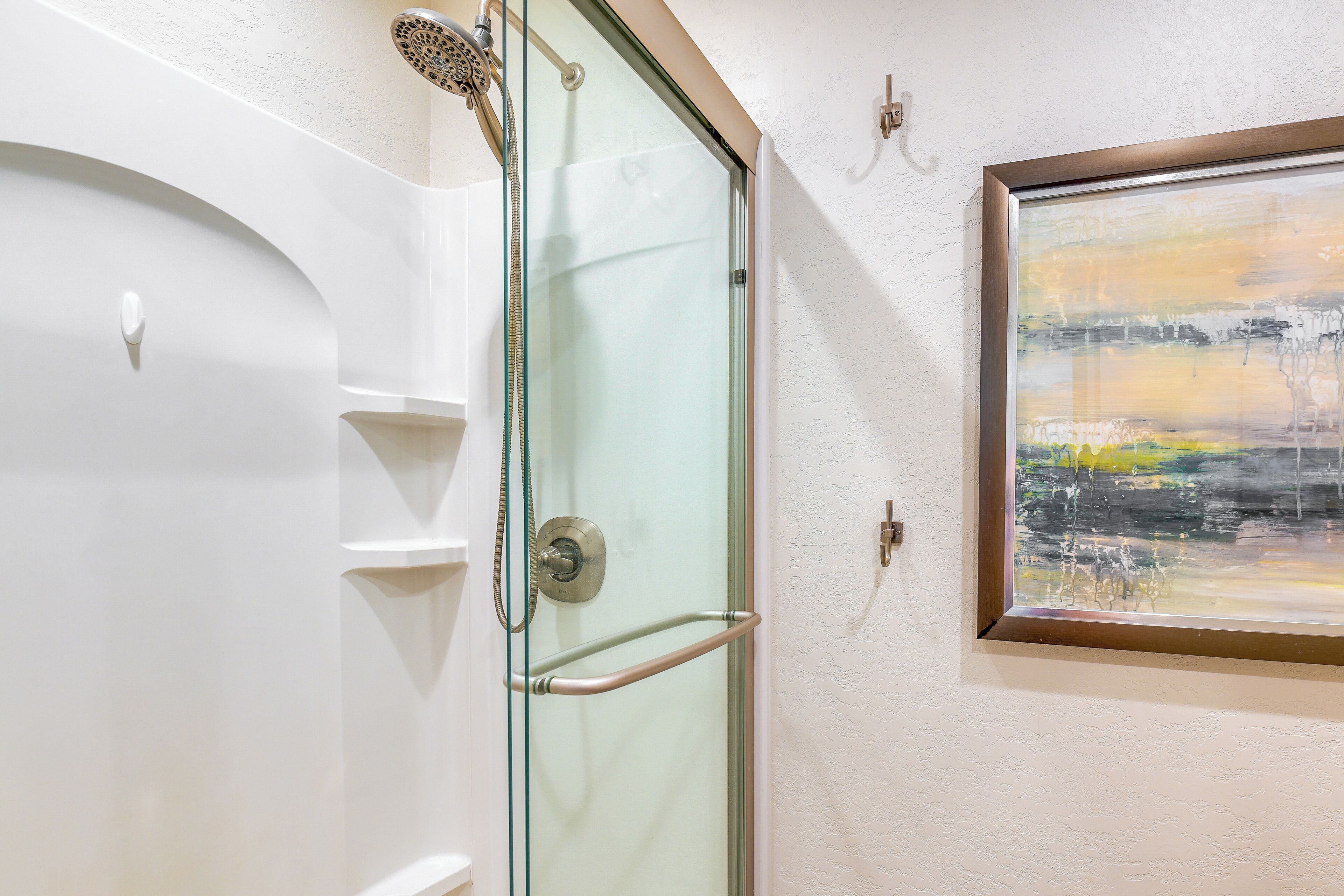 A glass-enclosed shower with a handrail, a white wall, and a framed abstract painting.