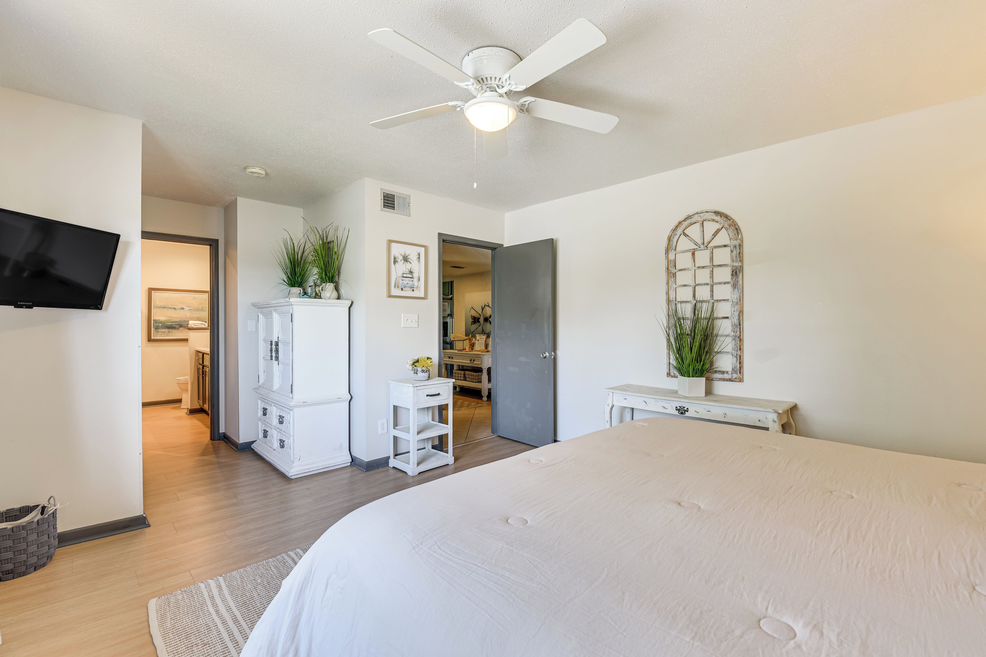 A modern bedroom with a large bed, a ceiling fan, a flat-screen TV, and a view into the bathroom.