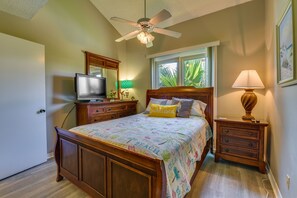 A bedroom with a wooden bed, a ceiling fan, a TV, a nightstand with a lamp, and a window with a view of palm trees. - Miramar Beach Rental w/ Pool Access & Golf Course! (Miramar Beach)