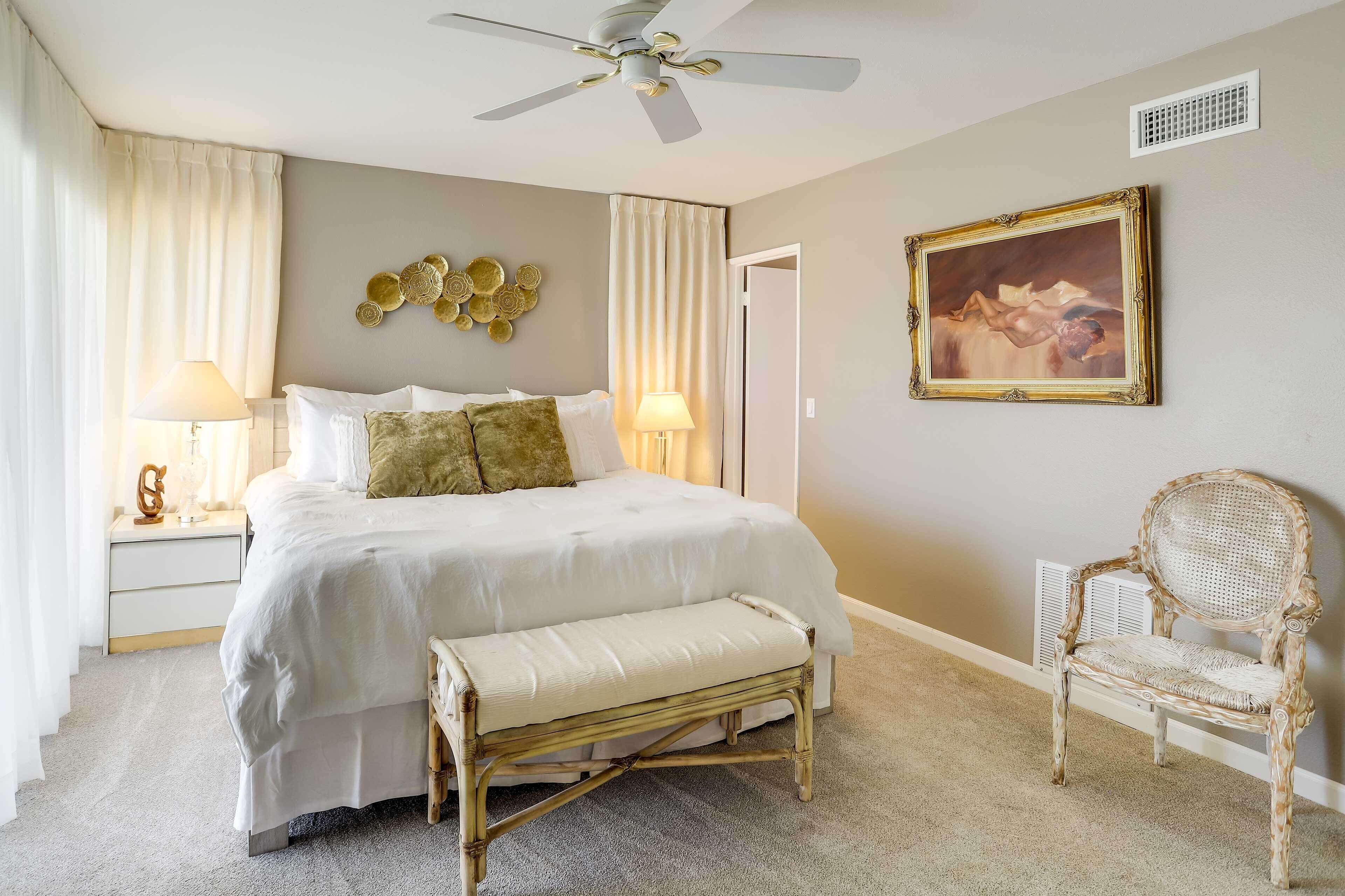 A bedroom with a bed, bedside lamps, a bench, a chair, a ceiling fan, a painting, and a vent.