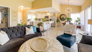 Ein modernes Wohnzimmer mit einem dunkelgrauen Sofa, einem runden Couchtisch und einem blauen Sessel. Im Hintergrund ist eine Küche mit weißen Schränken und Edelstahlgeräten zu sehen.