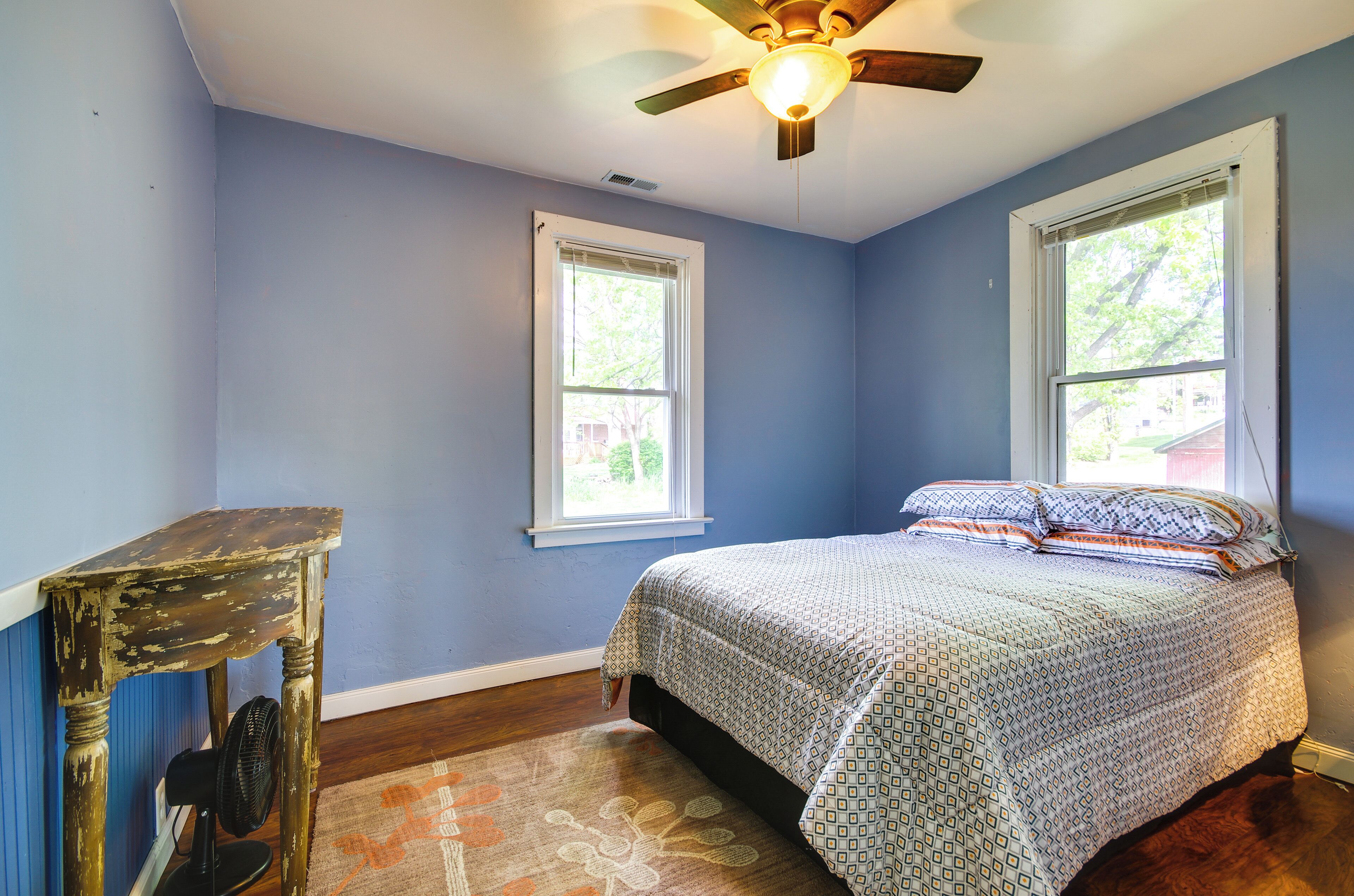 A bedroom with a bed, two windows, a ceiling fan, and a wooden table.