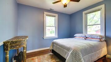 A bedroom with a bed, two windows, a ceiling fan, and a wooden table.