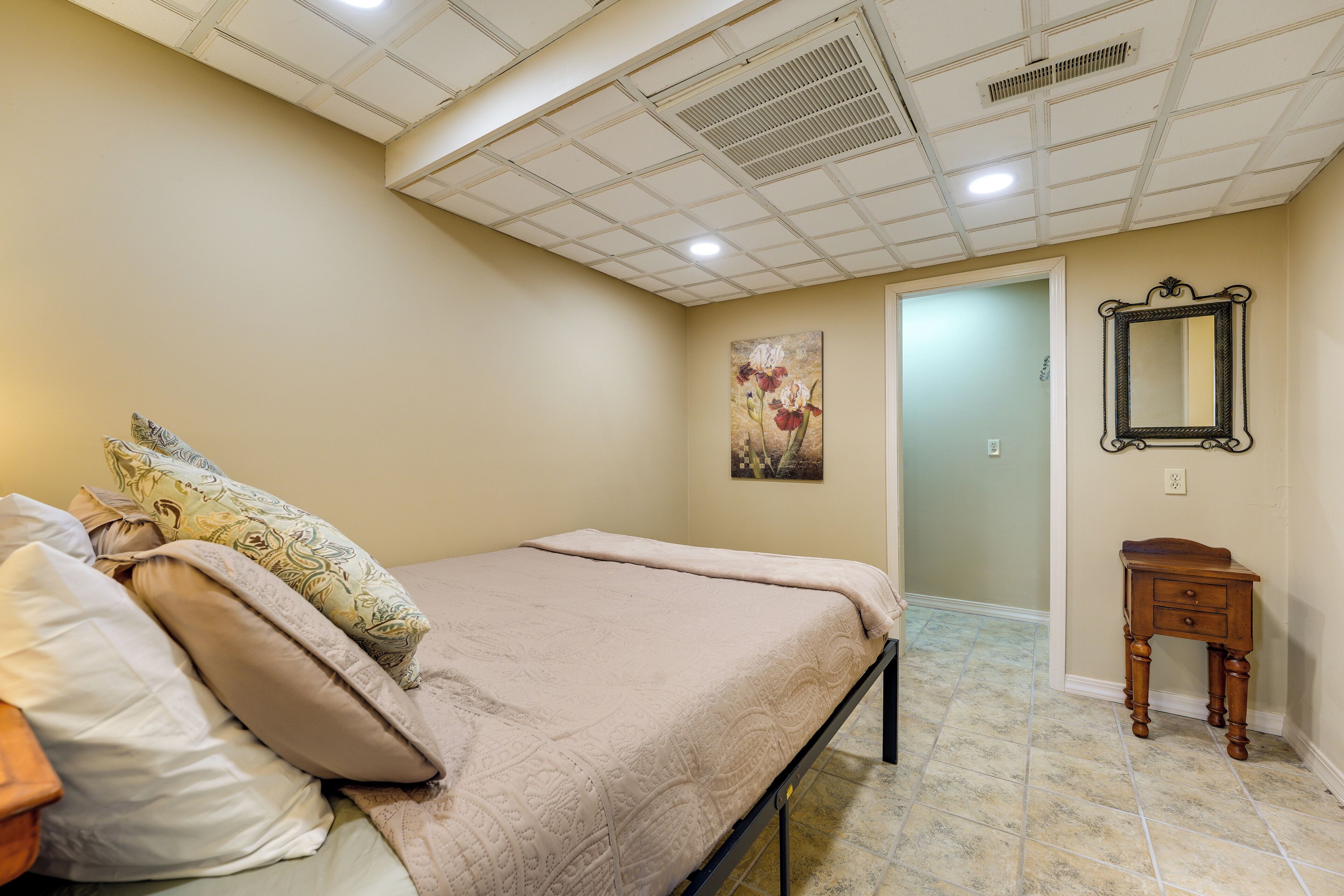 Quarto com cama, travesseiros, mesa de cabeceira, quadro, espelho e porta que dá para outro cômodo.