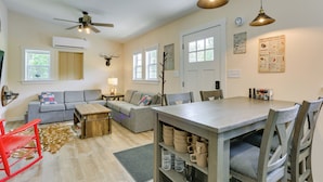 Ein geräumiges Wohnzimmer mit grauem Sofa, einem Holztisch und einem Esstisch mit Stühlen. Es gibt einen Deckenventilator, eine Wanduhr und ein Poster an der Wand.