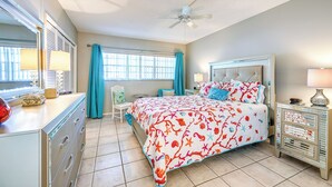 Ein Schlafzimmer mit einem Bett, einem Schminktisch, einem Nachttisch, einem Deckenventilator und einem Fenster mit Vorhängen.