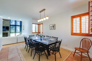 Un comedor con una mesa rectangular dispuesta para seis personas, rodeada de sillas negras. Se observa un amplio ventanal con marcos de madera a la derecha, y una chimenea con revestimiento de piedra a la izquierda.