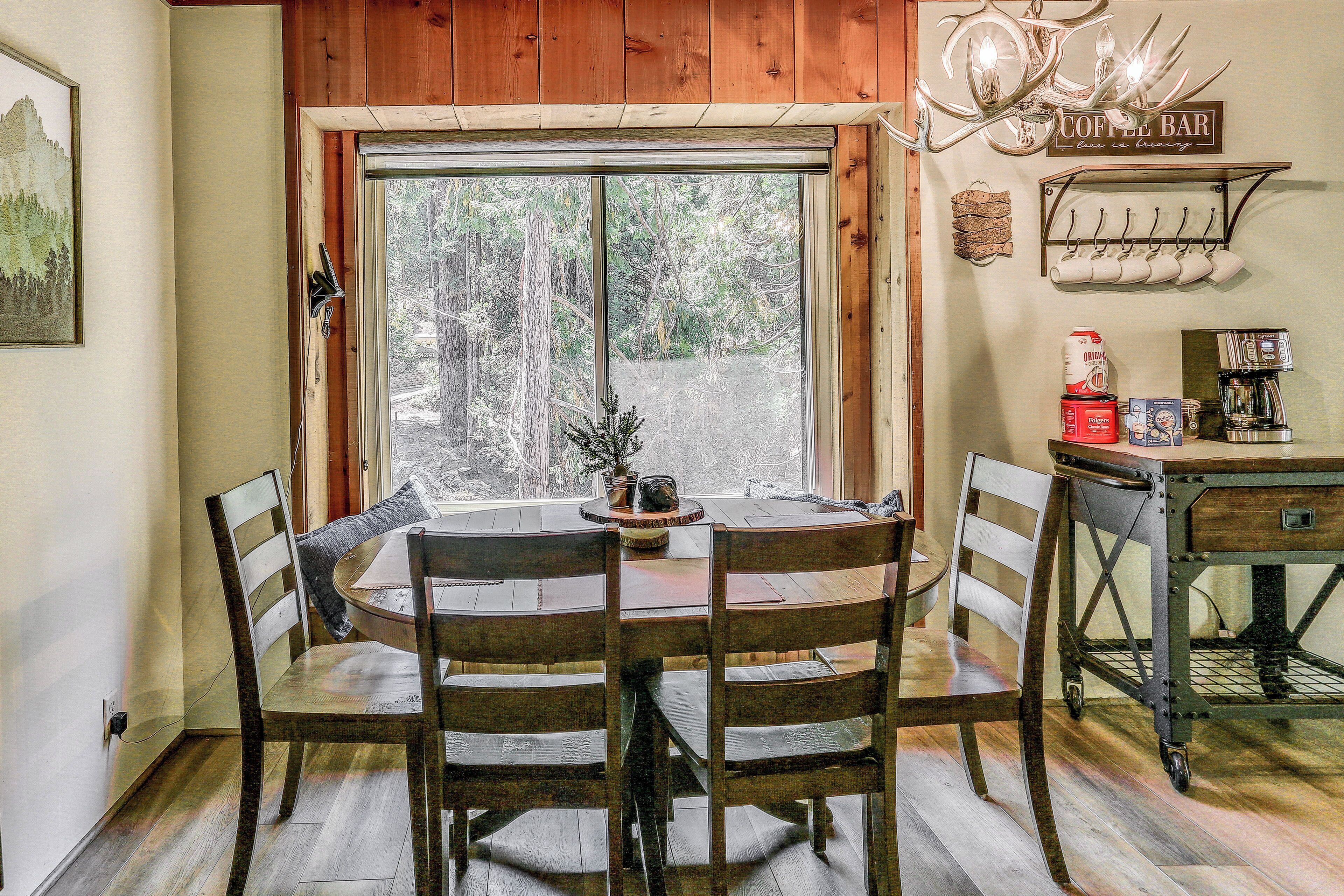 A dining area with a large window, wooden chairs, a round table, and a coffee bar with mugs and a coffee machine.