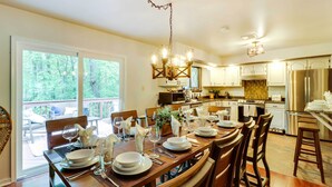Un comedor con una gran mesa de madera dispuesta para seis personas, rodeada de sillas. Un candelabro cuelga del techo, y hay ventanas que ofrecen vistas al exterior.