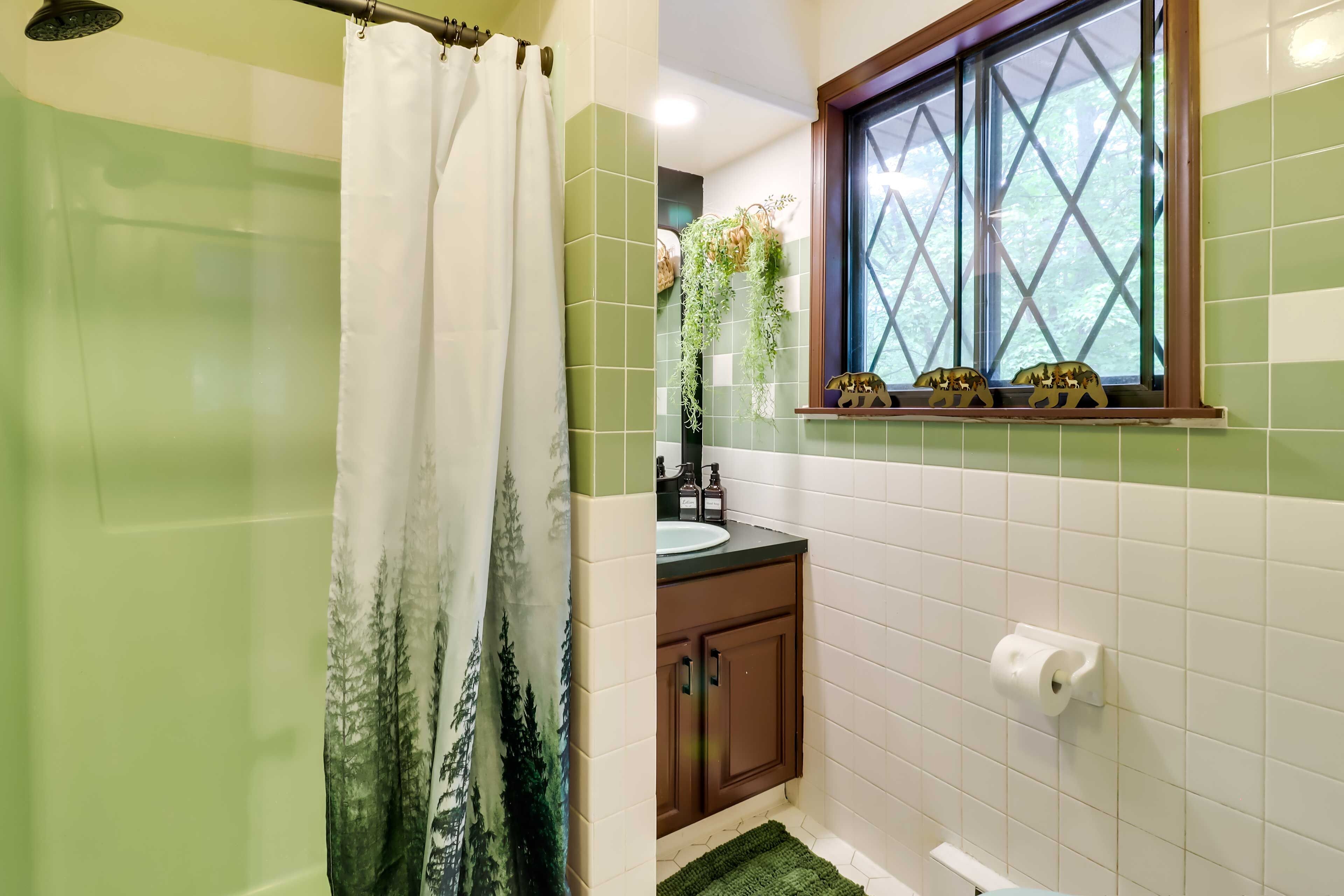 A bathroom with green tiles, a shower curtain with a forest design, a window with decorative items, and a toilet paper holder.