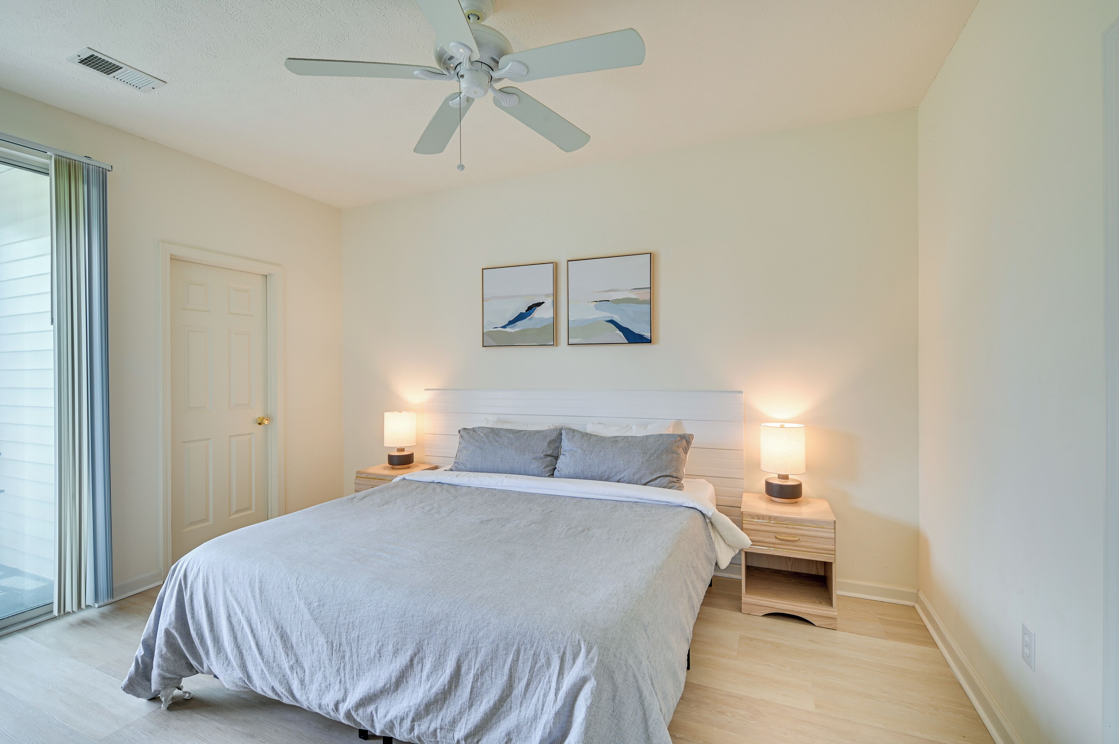Un dormitorio con cama, dos lámparas de noche, un ventilador de techo y dos cuadros en la pared.