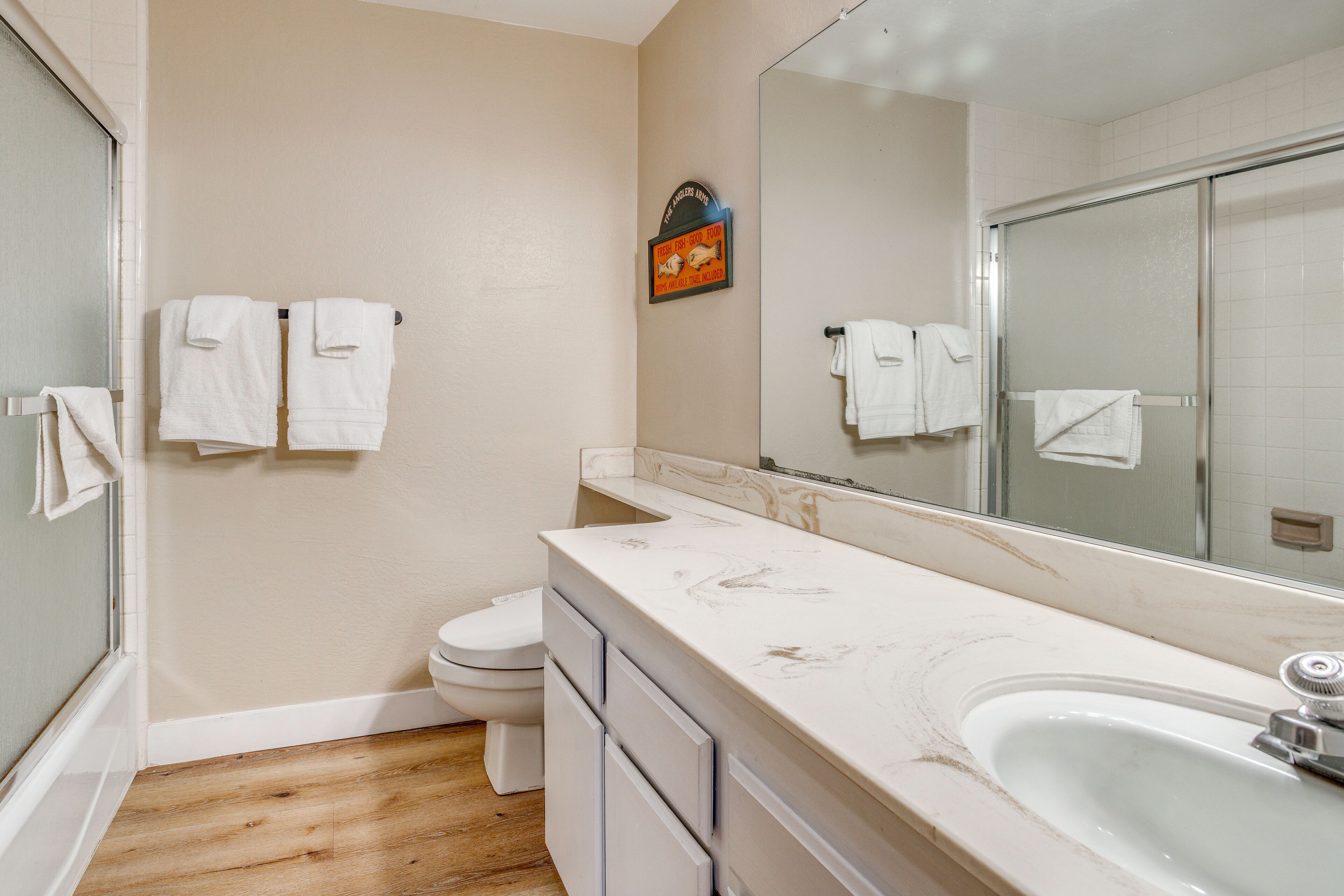 Un baño con piso de madera, lavamanos blanco, inodoro y ducha con puerta de cristal.