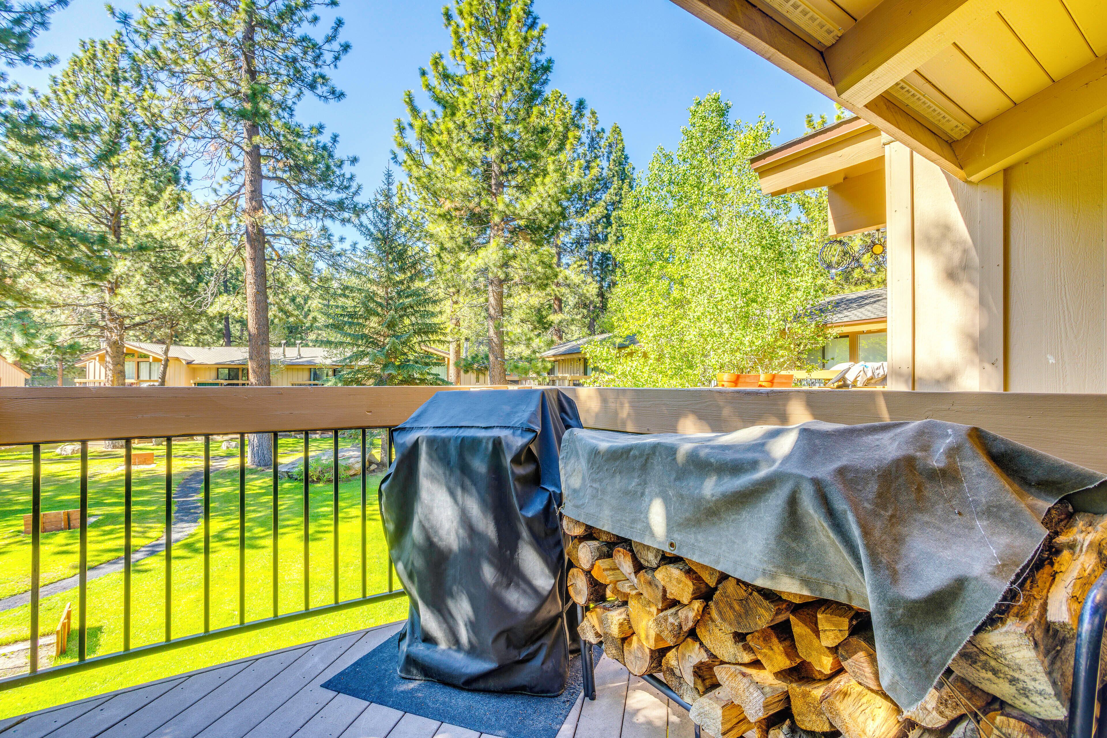 A deck with a grill and a covered firewood stack, surrounded by trees and a clear sky.